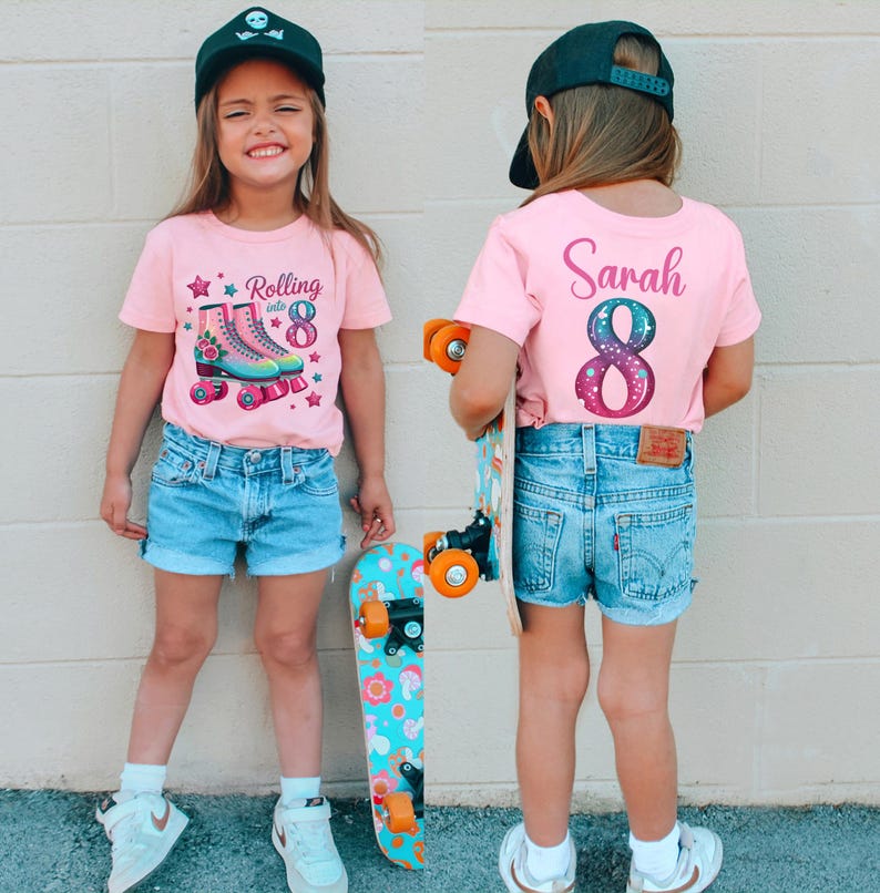 May include: A young person wearing a pink t-shirt with a roller skate graphic and the text "Rolling into 8". The person is also wearing denim shorts and a black baseball cap. A skateboard is held in the person's hand.