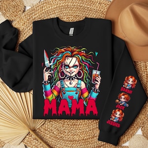 May include: Black sweatshirt featuring a colorful graphic of a doll-like character holding a knife and a drink, with the word "MAMA" in red. The sleeve displays three cartoon images with names: Sarah, Jack, and Jessica. A hat is visible in the background.