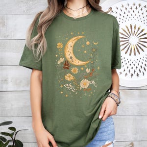 May include: A green t-shirt with a crescent moon and floral design. The moon is gold and the flowers are in shades of yellow, orange, and brown.