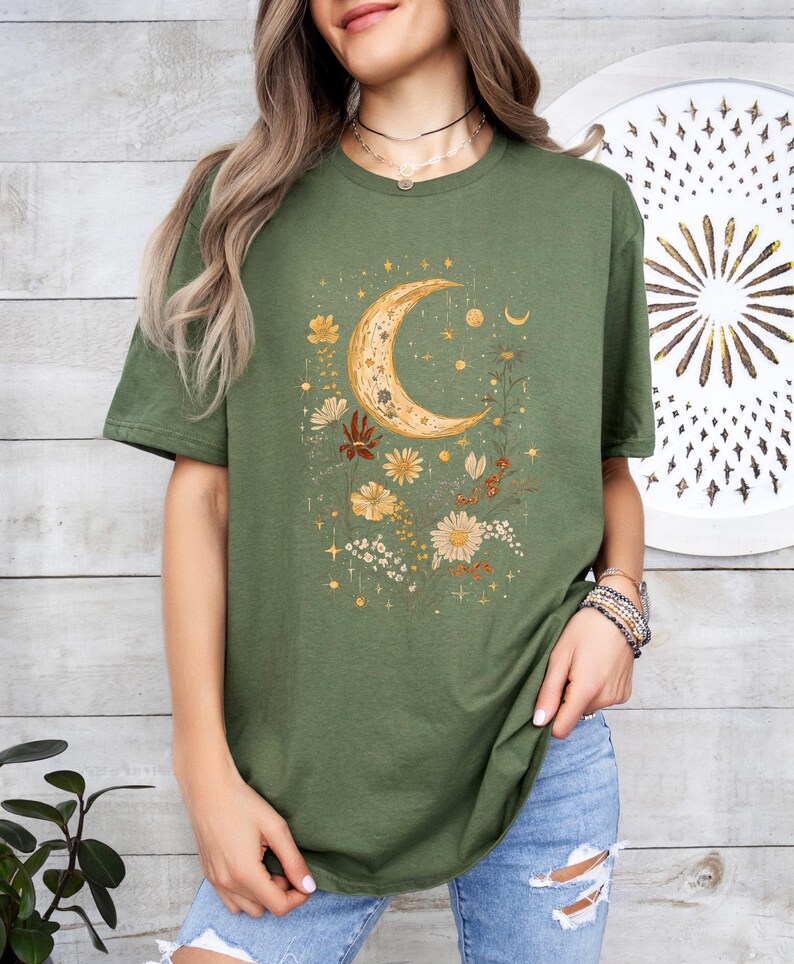 May include: A green t-shirt with a crescent moon and floral design. The moon is gold and the flowers are in shades of yellow, orange, and brown.