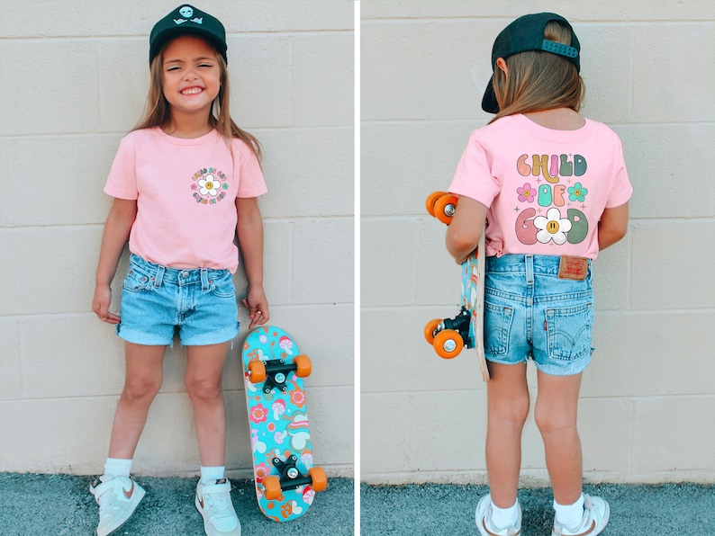 May include: A young person wearing a pink t-shirt with a flower graphic and the text "CHILD OF GOD" on the back. They are also wearing denim shorts, a black baseball cap, and white sneakers. They are holding a colorful skateboard.