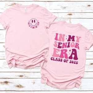 Puede incluir: Camiseta rosa con un gr&aacute;fico de cara sonriente en la parte delantera y el texto "In My Senior Era Class of 2025" en la parte posterior.