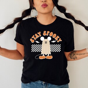 May include: Black t-shirt with the words "STAY SPOOKY" in orange, featuring a cartoon ghost character with a mouse-like head, orange shoes, and a checkered flag design. The shirt is a Halloween-themed graphic tee.