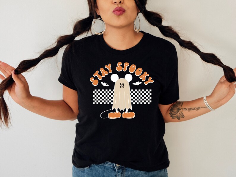May include: Black t-shirt with the words "STAY SPOOKY" in orange, featuring a cartoon ghost character with a mouse-like head, orange shoes, and a checkered flag design. The shirt is a Halloween-themed graphic tee.