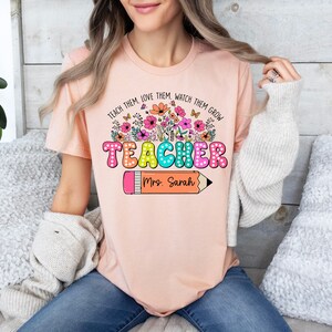 May include: A pink t-shirt with the text "Teach them, love them, watch them grow" above the word "Teacher" in colorful letters with white polka dots. A pencil with the text "Mrs. Sarah" is below the word "Teacher".