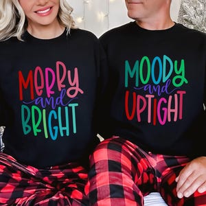 May include: Black sweatshirts with colorful text that reads "Merry and Bright" and "Moody and Uptight." The subjects are wearing red and black plaid pajama pants. The image is festive and holiday-themed.