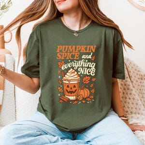 Może przedstawiać: Zielona koszulka z oliwkowym nadrukiem z napisem "PUMPKIN SPICE and everything NICE" w kolorze pomarańczowym i białym. Grafika zawiera pumpkin spice latte, dynie i jesienne liście. Koszulka ma okrągły dekolt.