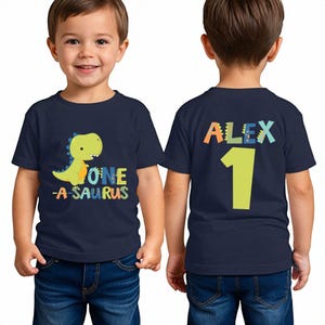 May include: Navy blue toddler t-shirt with a dinosaur graphic and the text "ONE -A-SAURUS" on the front. The back of the shirt has the name "ALEX" and the number "1". The shirt is worn by a young child.
