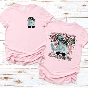 K&ouml;nnte beinhalten: Pinkes T-Shirt mit einem Motiv einer Frau, die eine blaue Baseballkappe mit einem Smiley und Sonnenbrille tr&auml;gt. Der Text "Mom Life All Day Every Day" ist auf der R&uuml;ckseite des Shirts aufgedruckt.