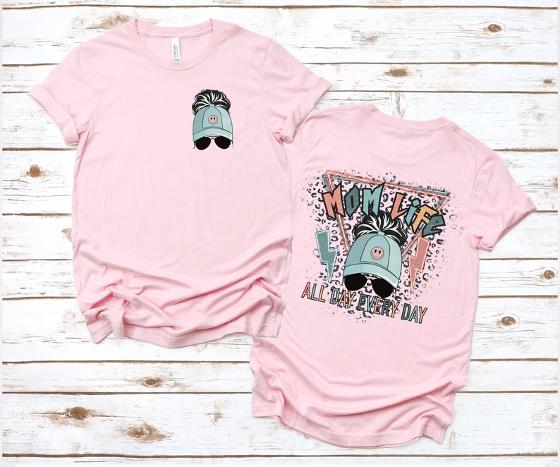 K&ouml;nnte beinhalten: Pinkes T-Shirt mit einem Motiv einer Frau, die eine blaue Baseballkappe mit einem Smiley und Sonnenbrille tr&auml;gt. Der Text "Mom Life All Day Every Day" ist auf der R&uuml;ckseite des Shirts aufgedruckt.