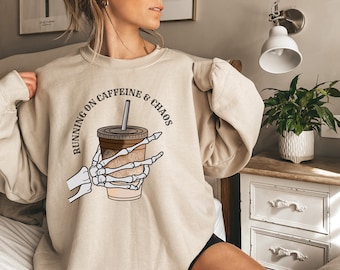 May include: A beige sweatshirt with a graphic of a skeleton hand holding a cup of coffee. The text "Running on Caffeine & Chaos" is printed in a circle around the graphic.