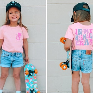 May include: A young person wearing a pink t-shirt with a mermaid graphic on the front and a black skull and crossbones cap. The person is holding a skateboard with a colorful design. The back of the t-shirt has the text "In My Mermaid Era" in pink, blue, and purple.