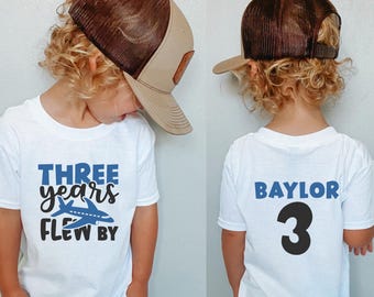 May include: A white t-shirt with blue text that says "THREE years FLEW BY" and a blue airplane graphic. The back of the shirt has blue text that says "BAYLOR 3". A child is wearing the shirt and a brown and beige baseball cap.