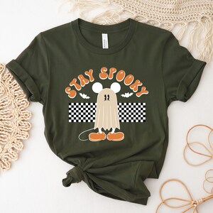 May include: Dark green t-shirt with the words "STAY SPOOKY" in orange lettering. The design features a cartoon ghost with mouse ears and orange shoes, a bat, and a black and white checkered pattern.