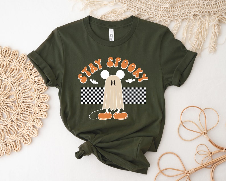 May include: Dark green t-shirt with the words "STAY SPOOKY" in orange lettering. The design features a cartoon ghost with mouse ears and orange shoes, a bat, and a black and white checkered pattern.