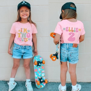 May include: A pink t-shirt with the text "I'm Two Cute" in colorful letters. The shirt is being worn by a child who is holding a skateboard. The child is also wearing a black baseball cap and denim shorts.