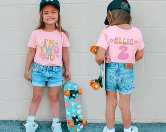 May include: A pink t-shirt with the text "I'm Two Cute" in colorful letters. The shirt is being worn by a child who is holding a skateboard. The child is also wearing a black baseball cap and denim shorts.