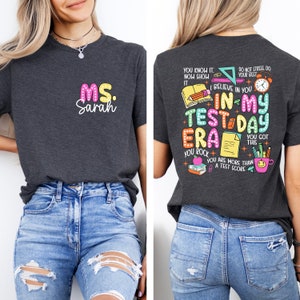 May include: A dark gray t-shirt with the text "MS. Sarah" on the front and a colorful graphic on the back that says "You know it. Now show it. I believe in you. In my test-a-day era. You rock. You got this. You are more than a test score."