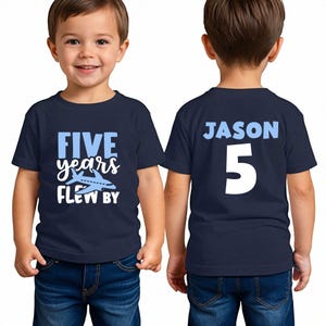 May include: Navy blue t-shirt with the text "FIVE years FLEW BY" and an airplane graphic. The back of the shirt has the name "JASON" and the number "5" in white. The child is wearing blue jeans.