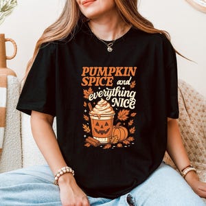Może przedstawiać: Czarny t-shirt z napisem "PUMPKIN SPICE and everything NICE" w kolorze pomarańczowym i białym. Grafika zawiera pumpkin spice latte, dynie i jesienne liście. T-shirt z grafiką w tematyce jesiennej.