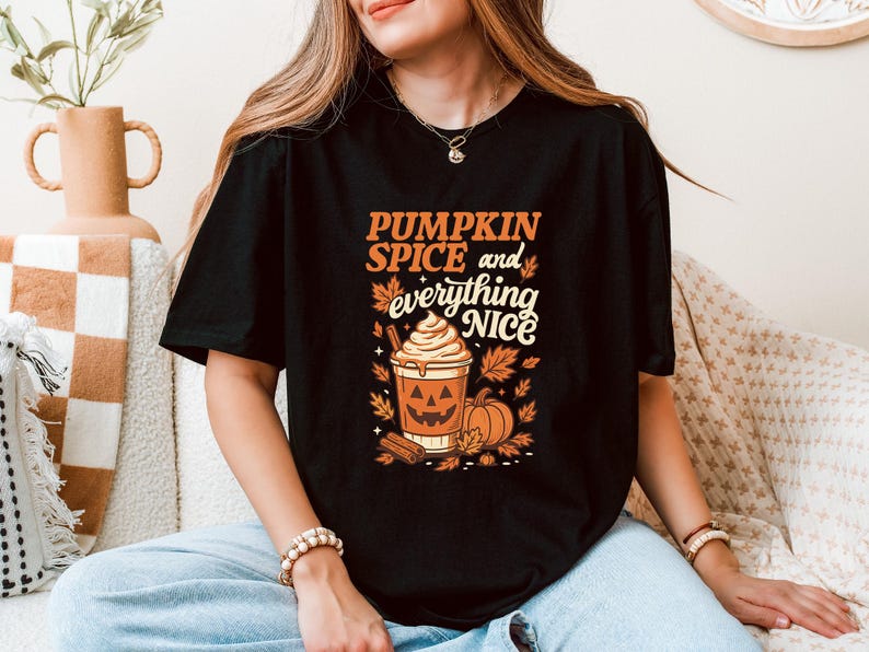 Może przedstawiać: Czarny t-shirt z napisem "PUMPKIN SPICE and everything NICE" w kolorze pomarańczowym i białym. Grafika zawiera pumpkin spice latte, dynie i jesienne liście. T-shirt z grafiką w tematyce jesiennej.