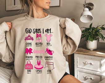 May include: A beige sweatshirt with the text "God Says I Am..." and pink illustrations of a crown, ballet shoes, a leotard, a bow, a dance bag, a tutu, a ballet barre, and scripture verses.