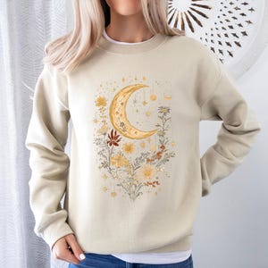 May include: A beige sweatshirt with a crescent moon and floral design. The moon is gold and the flowers are in shades of yellow, orange, and red.