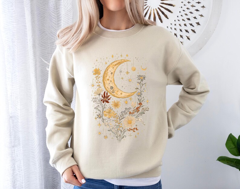 May include: A beige sweatshirt with a crescent moon and floral design. The moon is gold and the flowers are in shades of yellow, orange, and red.