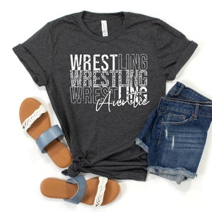 May include: A dark gray t-shirt with the text "Wrestling Auntie" in white. The text is stacked vertically with a distressed, glittery effect. The shirt is laid flat on a white background with a pair of blue and white sandals and denim shorts.