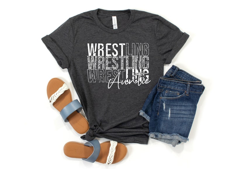 May include: A dark gray t-shirt with the text "Wrestling Auntie" in white. The text is stacked vertically with a distressed, glittery effect. The shirt is laid flat on a white background with a pair of blue and white sandals and denim shorts.