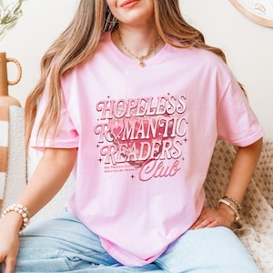 May include: A light pink t-shirt with the text "HOPELESS ROMANTIC READERS Club" in a retro font. The shirt also includes the text "WE PREFER OUR STORIES WRITTEN BY WOMEN" in a smaller font. The shirt is made of a soft material.