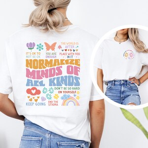May include: White t-shirt with a colorful graphic design that says "Normalize Minds of All Kinds". The graphic includes flowers, hearts, butterflies, a rainbow, and the text "It's OK to not be OK", "You are enough", "The world is after place with you in it", "Don't be so hard on yourself", "End the stigma", and "Keep going".
