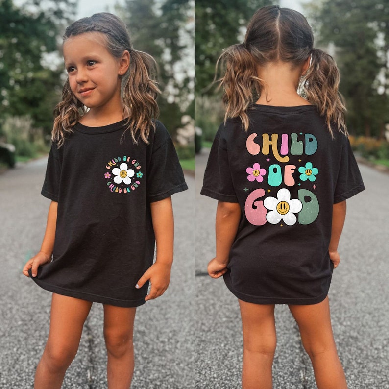 May include: A black t-shirt with a colorful graphic that says "Child of Good" with a daisy design. The shirt is being worn by a young person with long brown hair.