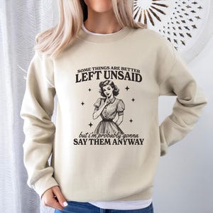 Puede incluir: Una sudadera beige con un gr&aacute;fico retro de una mujer con el dedo en los labios. El texto en la sudadera dice "Algunas cosas es mejor dejarlas sin decir, pero probablemente las diga de todos modos."