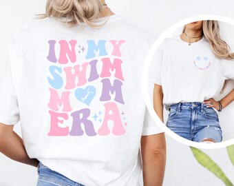 May include: White t-shirt with a pink, blue, and purple graphic that says "In My Swimmmer Era" on the back. The front of the shirt has a small pink and blue graphic of a smiling face with two hearts for eyes.