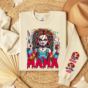 May include: Cream-colored sweatshirt featuring a colorful graphic of a doll holding a knife and a drink, with the word "MAMA" in red. The sleeve has cartoon images of two dolls with the names "SARAH" and "JACK".