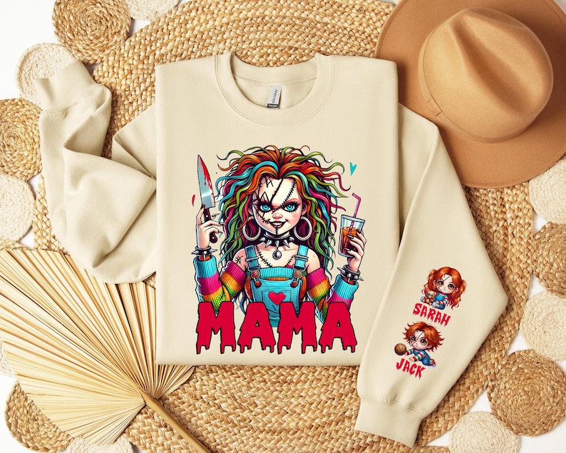 May include: Cream-colored sweatshirt featuring a colorful graphic of a doll holding a knife and a drink, with the word "MAMA" in red. The sleeve has cartoon images of two dolls with the names "SARAH" and "JACK".
