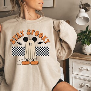 May include: Beige sweatshirt with a Halloween-themed graphic. The design features a ghost character with Mickey Mouse ears, orange shoes, and the words "Stay Spooky" in orange lettering. Two black bats are also included in the design.