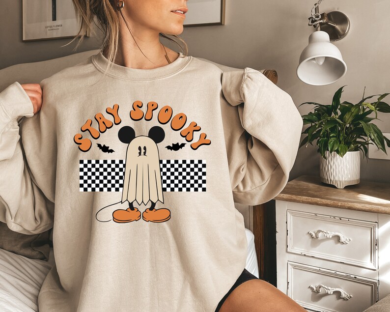 May include: Beige sweatshirt with a Halloween-themed graphic. The design features a ghost character with Mickey Mouse ears, orange shoes, and the words "Stay Spooky" in orange lettering. Two black bats are also included in the design.