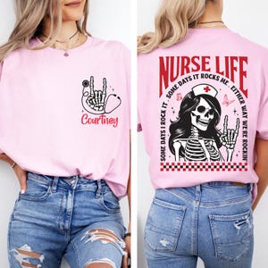 May include: A light pink t-shirt with a graphic design for nurses. The front features a skeleton hand with a stethoscope and the name "Courtney". The back of the shirt has the words "NURSE LIFE" and a skeleton nurse graphic.