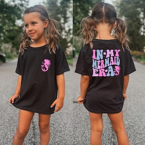 May include: A black t-shirt with a pink mermaid graphic on the front and the text "IN MY MERMAID ERA" in pink and blue on the back.