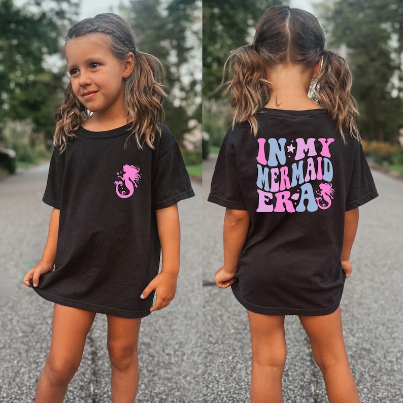 May include: A black t-shirt with a pink mermaid graphic on the front and the text "IN MY MERMAID ERA" in pink and blue on the back.