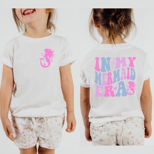 May include: White t-shirt with a pink mermaid silhouette on the front and the text "IN MY MERMAID ERA" in pink, blue, and purple on the back.