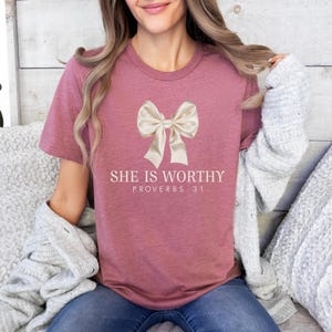 May include: A mauve t-shirt with a large white bow and the text "She is Worthy Proverbs 31" printed on the front.