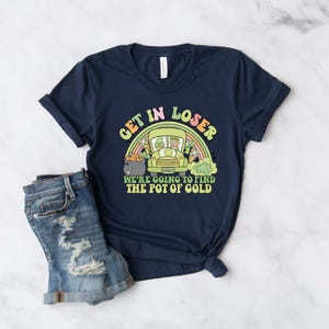 May include: Navy blue t-shirt with the text "GET IN LOSER" above an illustration of a green vehicle with leprechauns and a rainbow. The text "WE'RE GOING TO FIND THE POT OF GOLD" is below the illustration. A pair of blue denim shorts is in the image.