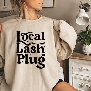 May include: A beige sweatshirt with the text "Local Lash Plug" in black, featuring stylized eyelashes and stars.