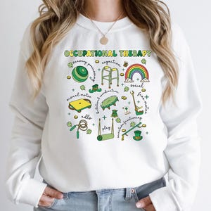 May include: White sweatshirt with the text "Occupational Therapy" and illustrations of various activities related to occupational therapy, including a rainbow, a walker, a ball, a jump rope, and a leprechaun hat.