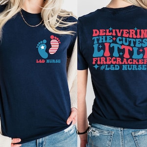 May include: Navy blue t-shirt with a red, white, and blue baby footprint design on the front and the text "L&D NURSE". The back of the shirt has the text "DELIVERING THE CUTEST LITTLE FIRECRACKERS #L&D NURSE" in red, white, and blue.