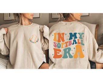 May include: A beige sweatshirt with a softball graphic on the front and the text "In My Softball Era" in colorful retro lettering on the back.