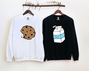 May include: Two white and black sweatshirts with a cookie and a carton of milk graphic. The cookie is brown with chocolate chips and has a bite taken out of it. The milk carton is white with a blue lid and a straw sticking out of it. The text "MILK" is printed on the carton.